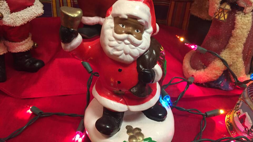 A small figurine of a Black Santa Claus sits on a table surrounded by multicolored string lights and other figurines.