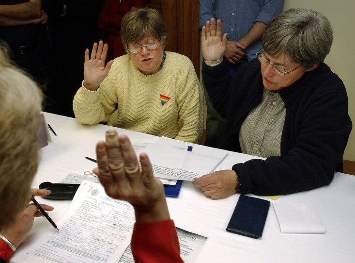 Two women raise their hands and recite an oath from a piece of paper, sitting across the table from an official also raising her hand. It's a florescent office.