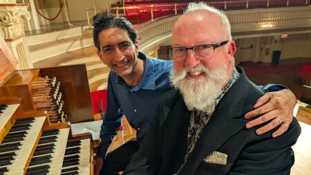 A man in a buttondown puts his arm around a man in a blazer. They're sitting together in front of an organ and smiling for the camera.