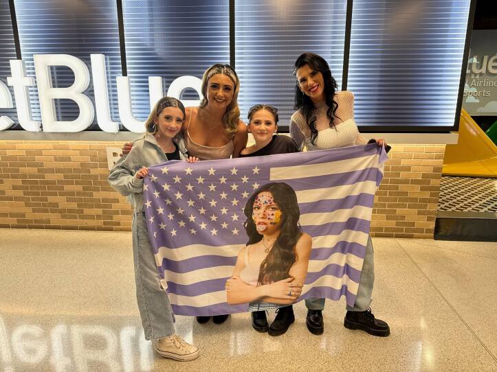 Two young girls and two women are holding up a purple flag in the style of the American flag, with a photo of singer Olivia Rodrigo on it.