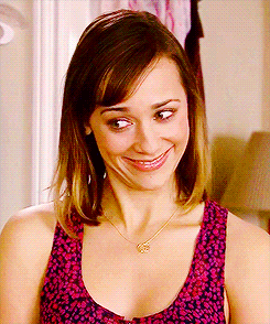 Rashida Jones, wearing a pink and blue speckled tank top, smiles awkwardly, raising her eyebrows.