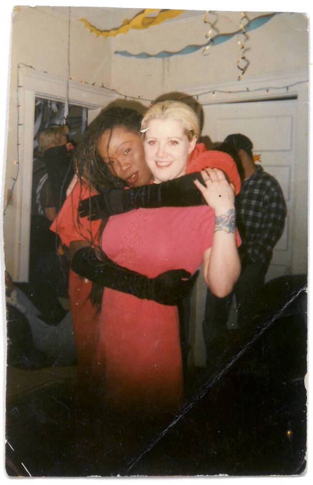 A woman hugs her friend from behind, arms wrapped around her shoulders and torso, as they both smile.