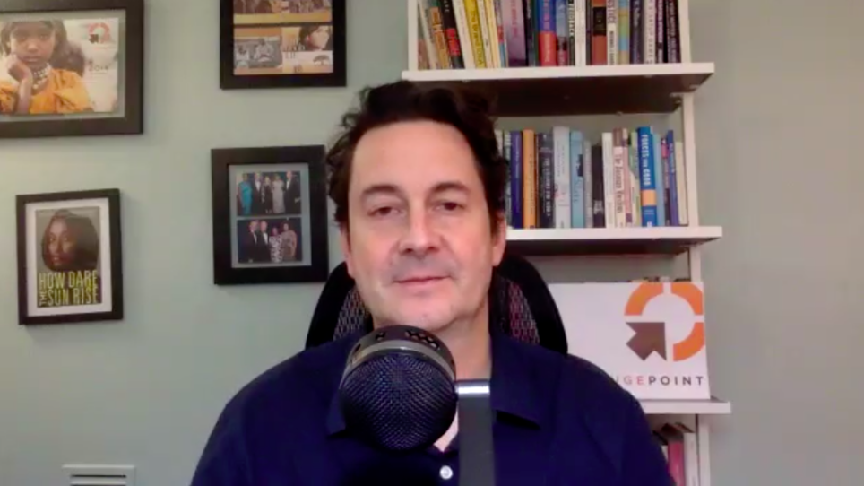 A man sits behind a microphone. Photographs and books are on the wall behind him.
