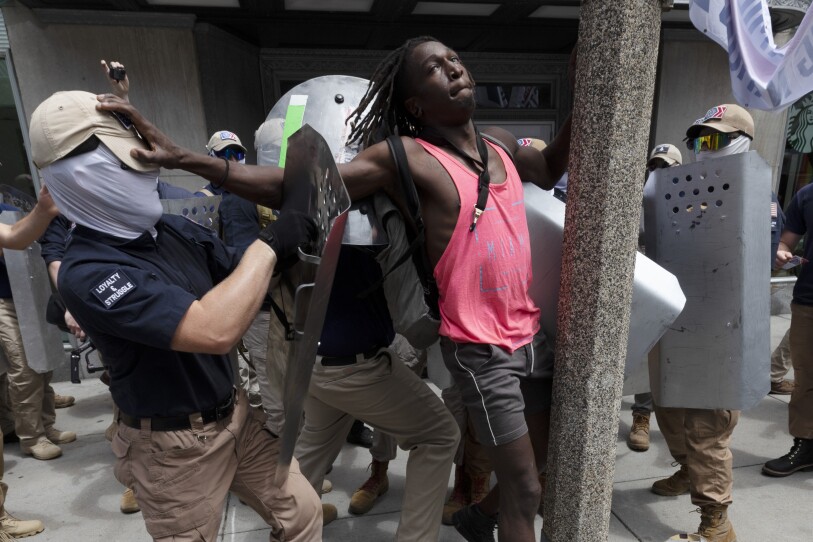 A group of masked men in matching clothes and face coverings use metal shields to push a Black man up against a post on the sidewalk.