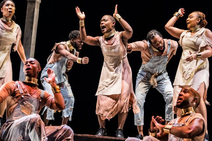 This is an image from a production of DRUMFOLK. A dance performance that is set in 1740, where enslaved Africans in South Carolina have been banned from using drums and other instruments. Instead of being silenced they create step dance, using their feet and hands to create percussive beats. In this image men and women are  onstage against a black background. They are step dancing step. Their hands are in the air as they clap and stomp. The men are wearing coveralls and a traditional African gold choker and gold cuffs on their wrists. The women are wearing tattered sheath dresses and traditional African gold chokers and bracelets.