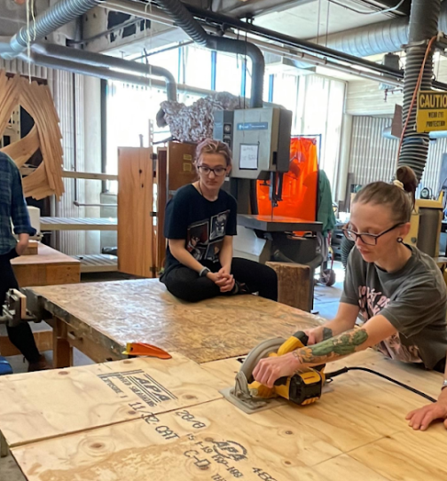 Two young people are in a workshop space, one uses a yellow table saw to cut plywood.