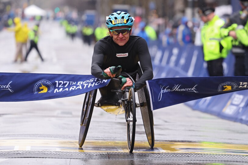 A woman in a racing wheelchair breaks the tape at the finish line. She wears a blue helmet. The pavement is wet.