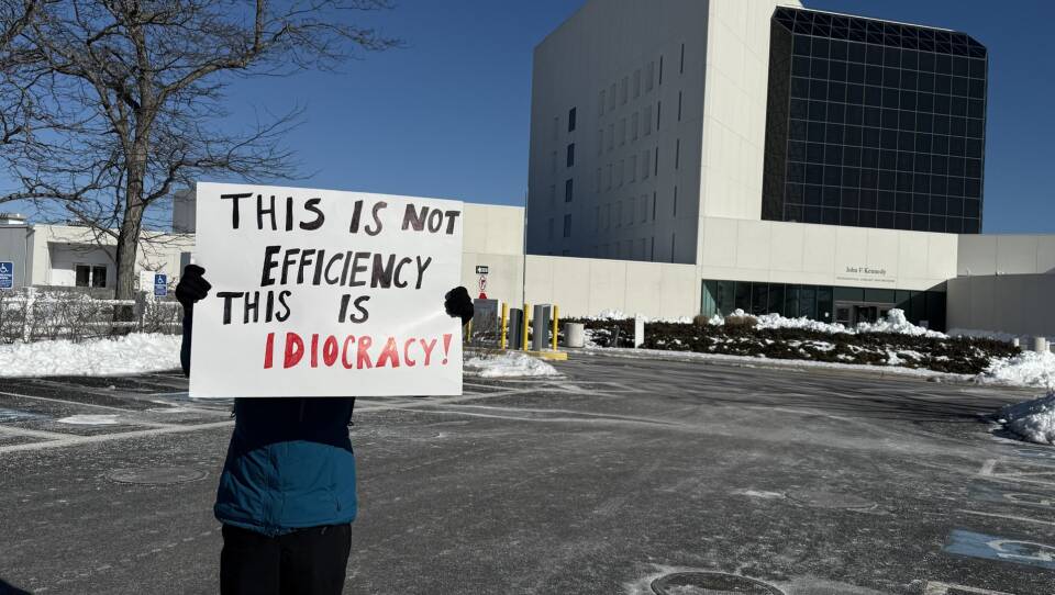 A person holding a sign in front of their face in front of the JFK Library & Museum that reads "this is not efficiency this is idiocracy."