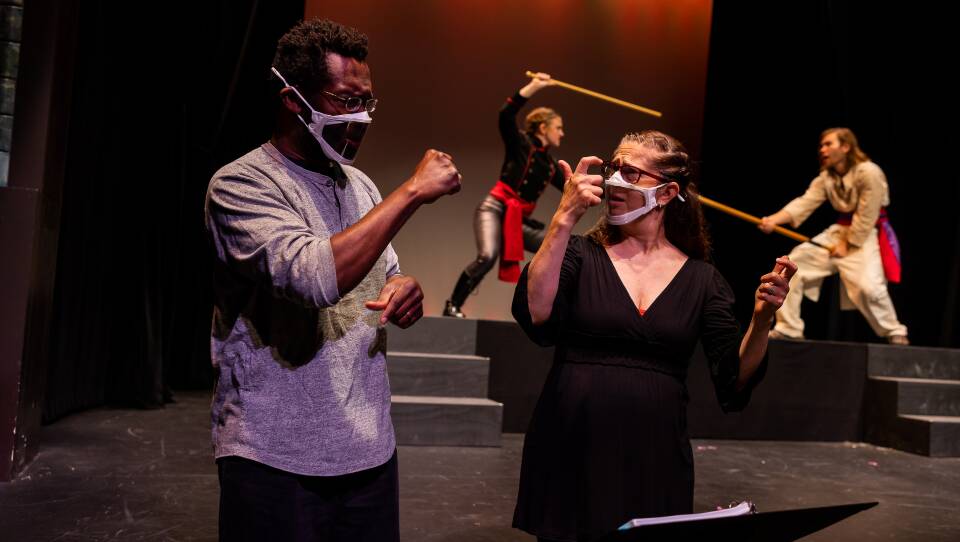 A man and woman in a mask speak to each other using sign language. Two actors on stage behind them engage in a sword fight.