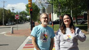 Two people laugh and smile, posing in front of a rainbow-painted crosswalk