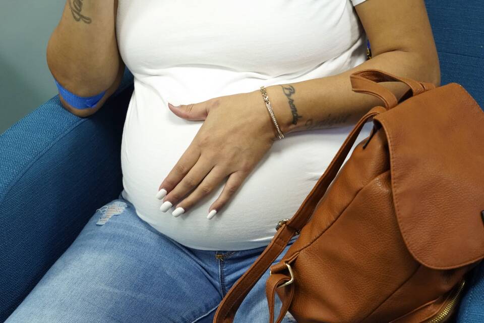 A pregnant woman rests one hand on her belly as she sits on a chair in a waiting room.