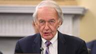 A man in a suit and tie sits behind a desk in a Capitol Hill hearing room.