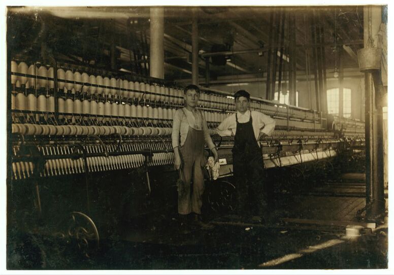 Two young children stand in a textile factory in this sepia photo