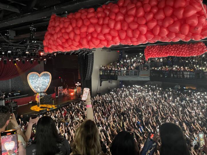 This is an interior photograph of a concert. Taken from a balcony, we see the main floor and hundreds of concertgoers holding their arms in the air. The crowd is lit with peoples' smart phones. A balloon drop appears to be imminent; along the ceiling are clear containers holding hundreds of red, heart shaped balloons.
