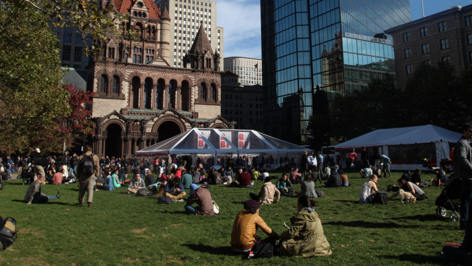 Dozens of people sit and walk on a grassy park in a city.