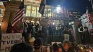 A crowd of people holding signs and flags stands in front wrought iron gates.