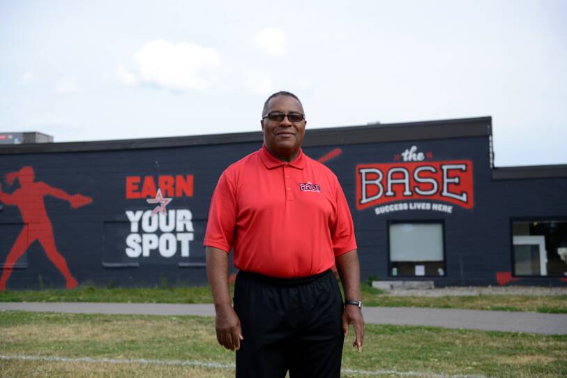 A man in a bright red polo shirt stands on a baseball field. Behind him, a building has a sign that reads "The BASE: success lives here"