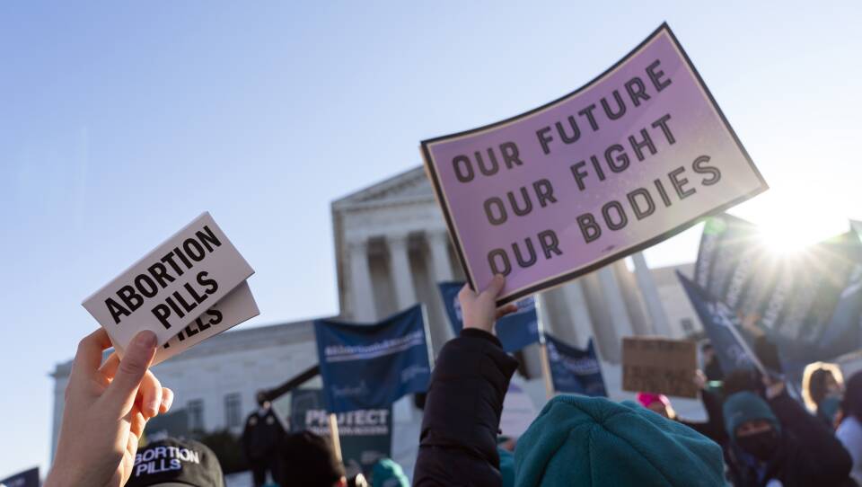 A crowd of protesters hold signs in front of the Supreme Court of the United States. Signs read "abortion pills" and "our future, our fight, our bodies"