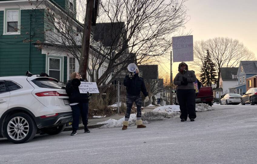 Three people, two holding anti-vaccine signs and one with a bullhorn, stand on a street with small snowbanks behind them.