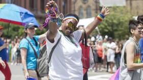 A person holds up rainbow beads and beams for the camera. A large crowd of onlookers are gathered in the background of the march.