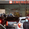 In the foreground, cars are clumped together, all with their brake lights on attempting to drive toward the emergency room. Outside the emergency room doors, more than a dozen people are lined up, wearing masks and appearing to be somewhat randomly clustered.