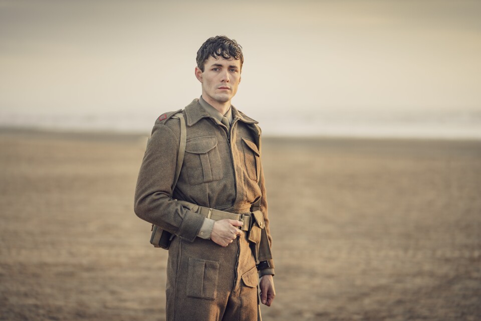 A young man stand in an empty, fallow field, and looks into camera. His hair is greasy and tousled, his face dirty, and he wears a British Soldier's WWII uniform.