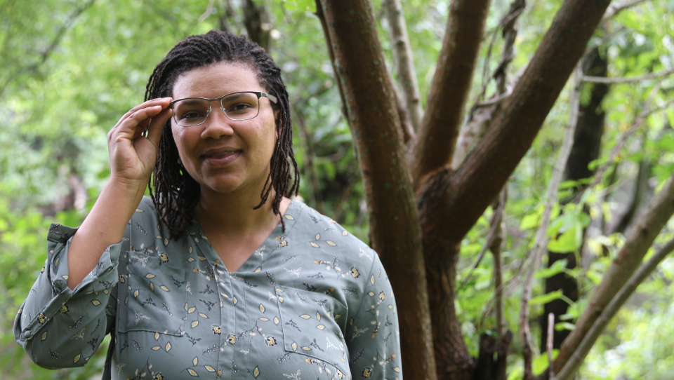 A woman, half-smiling, stands amid greenery and adjusts her glasses on an overcast day