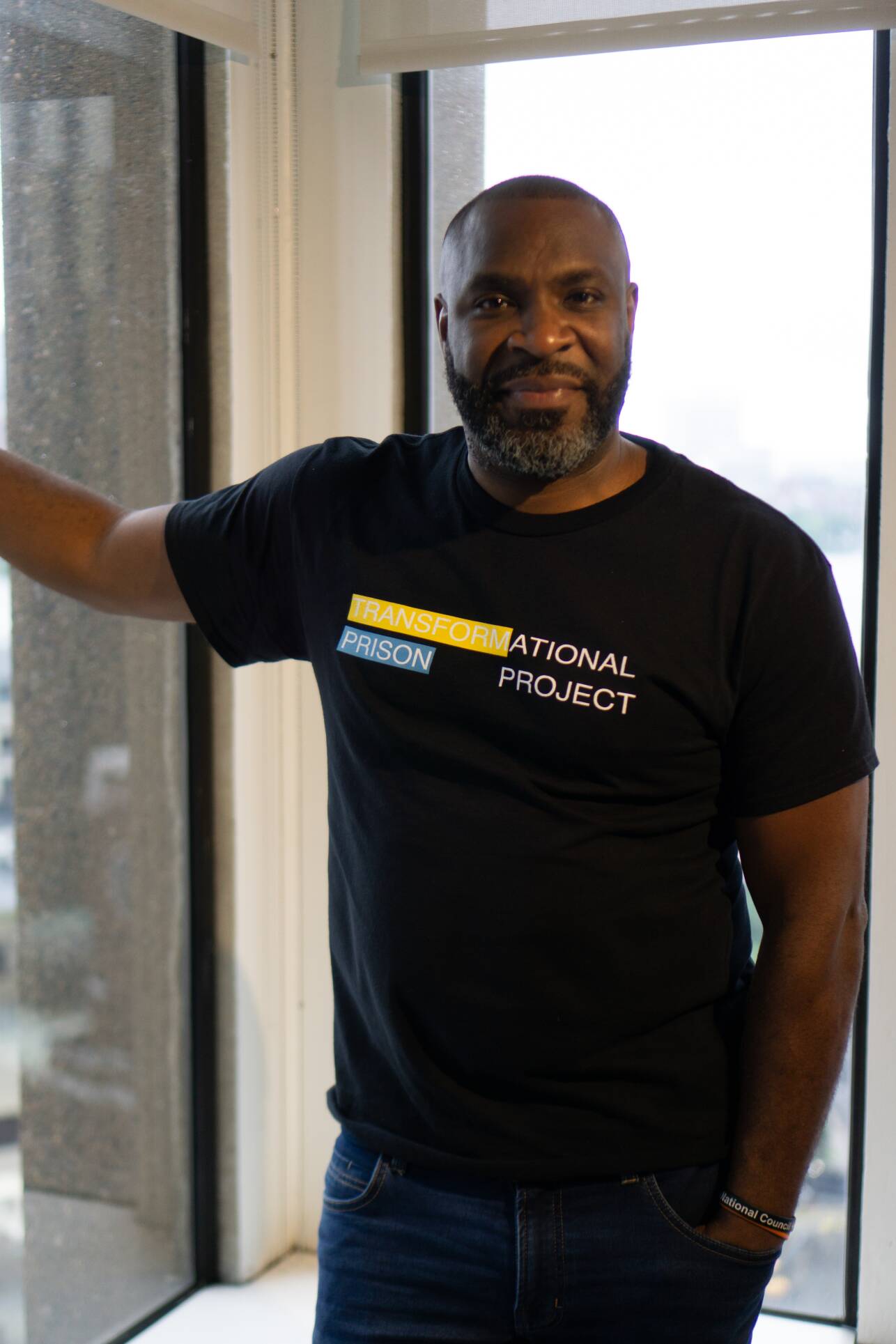 A man smiles for the camera in a black T-shirt that reads "Transformational Prison Project"