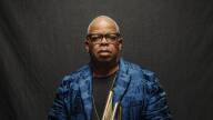 Terence Blanchard wears a blue velvet jacket and holds his trumpet against a black backdrop, a slight smile on his face.