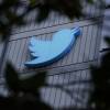 A baby-blue Twitter logo protrudes from the side of a building, photographed through tree branches