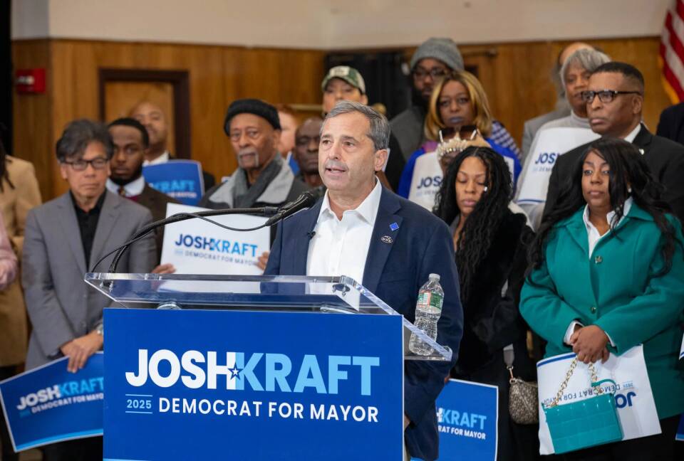 A man in a suit jacket speaks behind a lectern decorated with the sign "Josh Kraft Democrat for Mayor." Behind him are about a dozen people holding other signs.