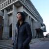 A woman with her hands in the pockets of her black jacket stands on the sidewalk outside a large concrete building.
