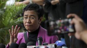 A woman in a pink jacket speaks before a group of reporter with microphones and cellphones.