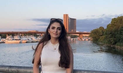 A woman stands and looks at the camera. Behind here is a harbor and the Encore Casino.