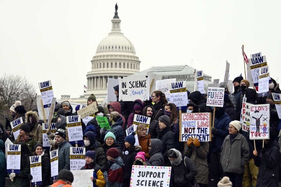 Dozens of scientists and advocates hold up signs with messages like "Support science" and "Congress makes laws not the president" in front of the White House.