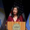 Michelle Wu speaks from behind a lectern on stage, with Boston city flags hanging behind her.