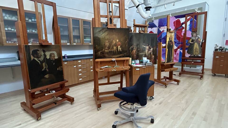 An empty swivel chair sits in front of a painting on a large easel in a workshop space, with other paintings stationed to both the left and right.