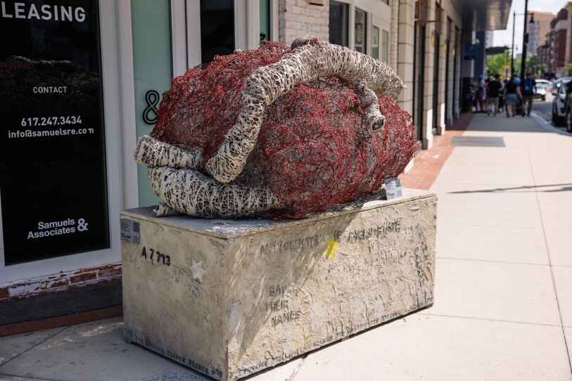 This is a photo of a public art sculpture, which is on the sidewalk. It's a rendition of a heart made from red and white chicken wire. It is mounted to a larger rectangle of concrete. It's titled “Prisoner a-7713.” "A-7713" is stenciled on the concrete. tihs is the number tattooed on Holocaust survivor Elie Wiesel’s arm when he arrived in Auschwitz.
