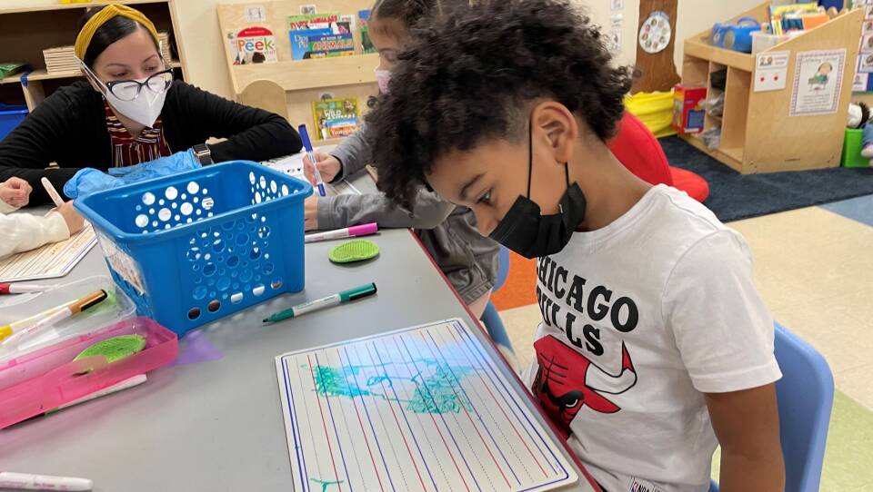 A woman wearing glasses and a mask kneels down at the end of a table where two young children and drawing on dry-erase boards.