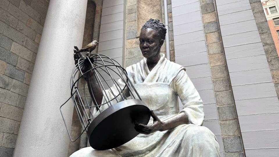 A massive bronze statue of a Black woman, seated and holding a birdcage that she's opened. A free bird sits on top of the bird cage, staring at the woman, who wears a simple white dress of stone or plaster.