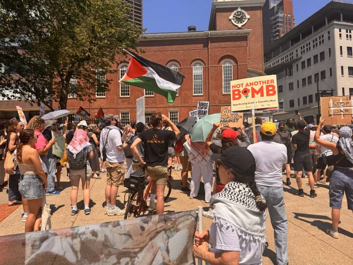 A group of people holding signs stands in front of the old State House building in Boston.