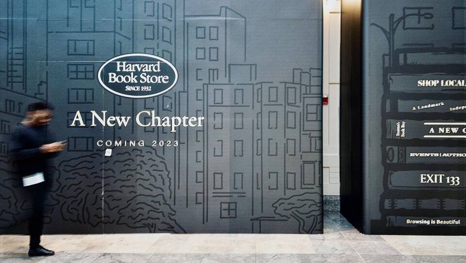 This is a photograph of the forthcoming Harvard Book Store at the Prudential Mall. The exterior of the storefront is covered in  black fabric with a rendering of a cityscape on it. Across this fabric it says "Harvard Book Store:  A New Chapter, Coming 2023" A person is seen walking past the book store