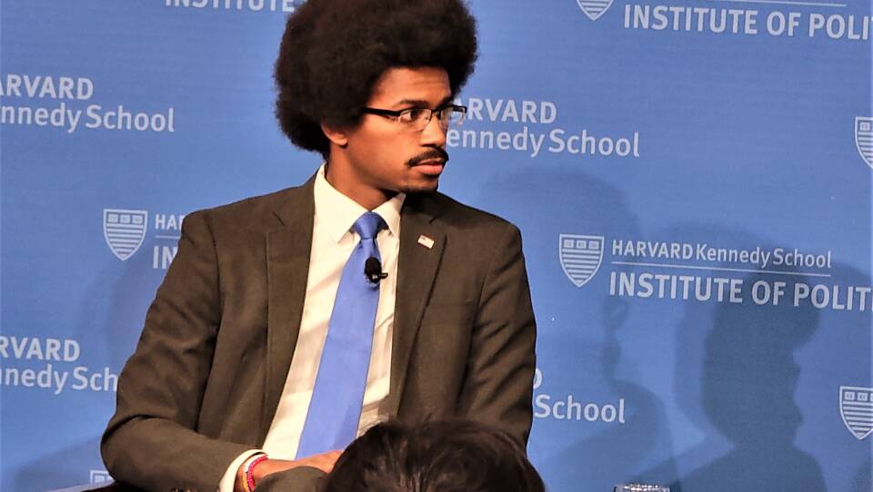 A man wearing a black suit and blue tie sits in a chair on a stage, with a background that reads "Harvard Kennedy School Institute of Politics.