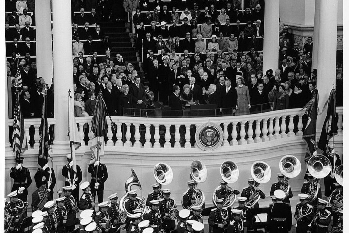 In a crowded grandstand full of guests and dignitaries, President Richard Nixon stands on stage with his right hand raised.