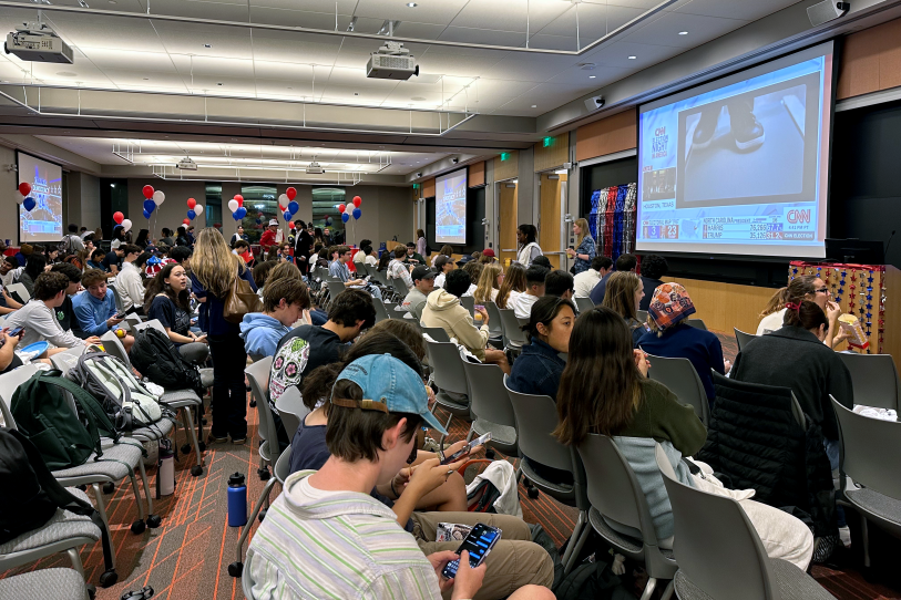 Students with backpacks sit in plastic chairs in what appears to be a lecture hall. CNN feeds are projected onto screens. Red white and blue decorations like balloons are around the room.