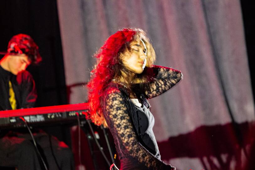 a girl sits on stage, touching her hair. In the background is a man on a piano.