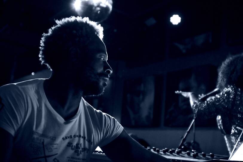 This is a black and white close up shot of keyboardist Ray Angry playing the keyboard during a live performance. This is a profile shot, we see only his upper body, in the background we see the back of a singer, who is facing the audience.