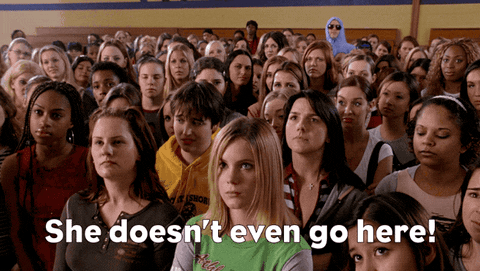 A man hiding in the back of a crowded room shouts "she doesn't even go here!"