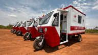 Five small red and white machines that read "Ambulance" are on motorized tricycles in a desert-like environment.