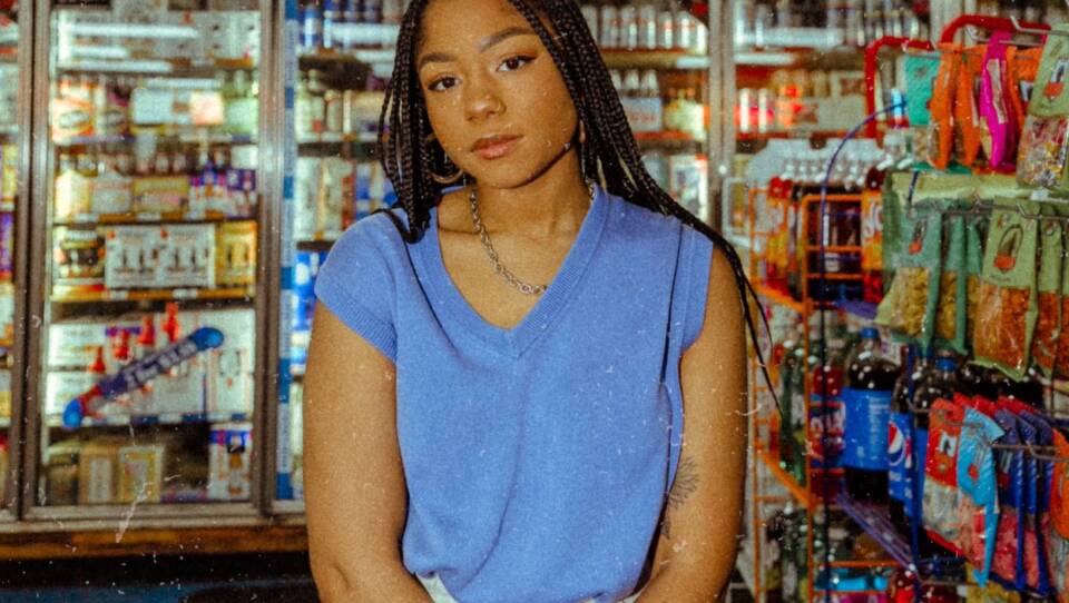 A young woman with braided hair looks toward the camera as she stands inside a convenience store.